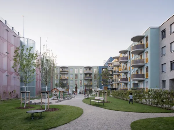 Außenanlagen mit Kinderspielplatz, spielenden Kindern und Bänken im Vordergrund. Diese werden umsäumt vom Parkhaus und vierstöckigen Wohngebäuden mit Balkonen und bunten Fassaden.