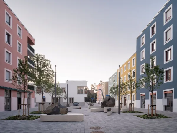 Blick vom mittleren Quartiersplatz auf die Kindertagesstätte. Rechts und links im Bild sieht man die bunten Wohngebäude. Im Vordergrund Sitzgelegenheiten aus Stein mit Kunst aus Stein und futuristische Außenbeleuchtung.